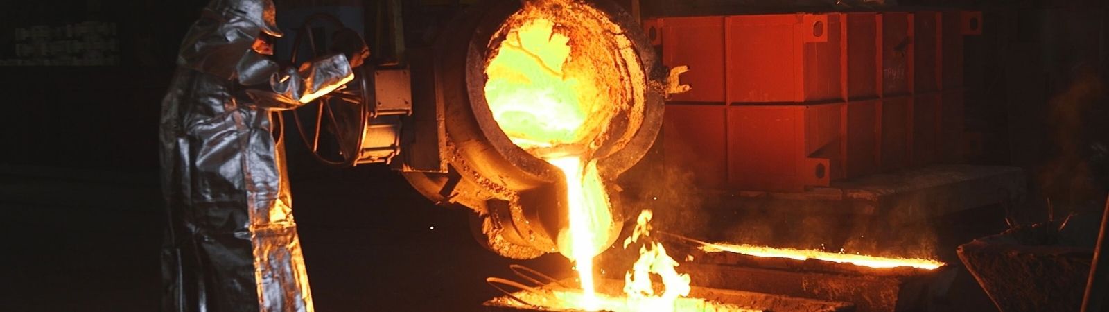 Smelter employee in a silver protective hazmat suit pouring molten gold from a heavy industrial pot into a container on the ground.