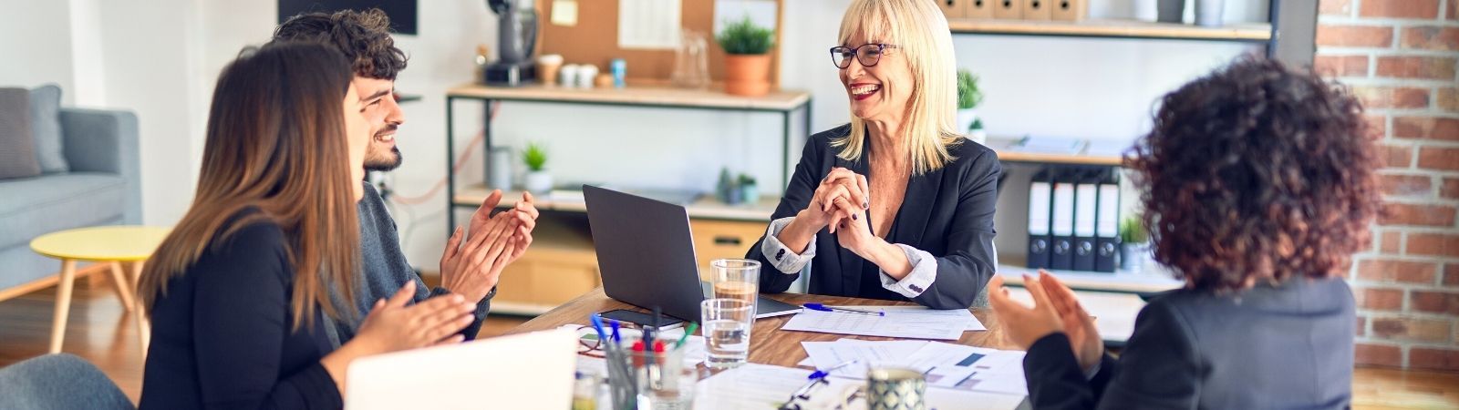 Three professional women and one man sitting around a table in a work meeting smiling and bouncing ideas off one another.