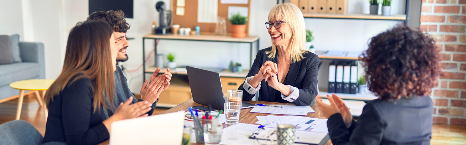 Three professional women and one man sitting around a table in a work meeting smiling and bouncing ideas off one another.