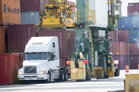 A white semi truck is parked at a terminal while cranes load containers.