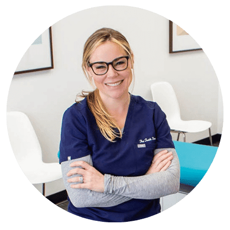 Dr. Megan Posthuma of The Tooth Ferry Pediatric Dentistry smiles in scrubs with folded arms, leaning against an exam table in her office