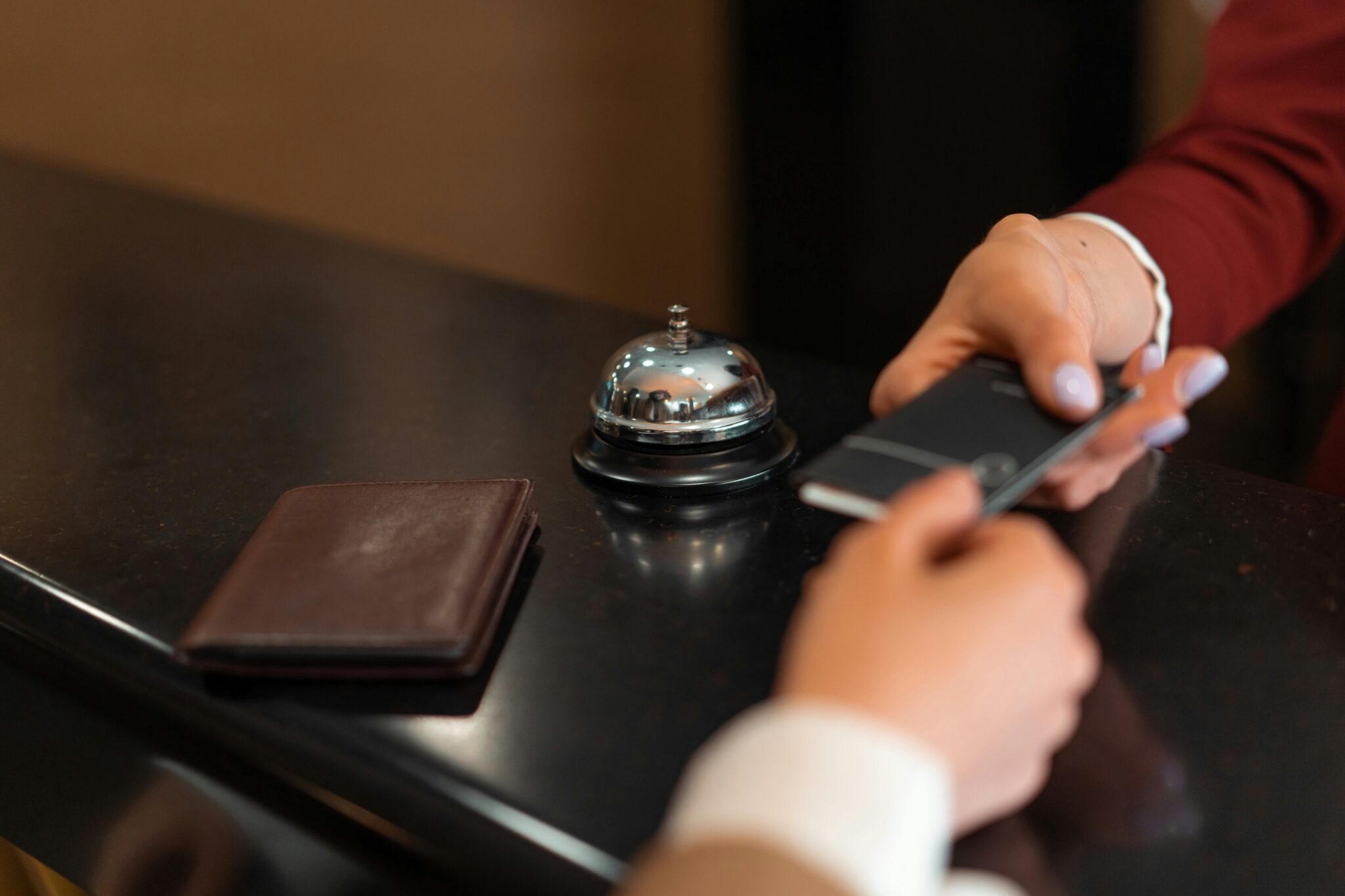 A person hands a smartphone to a receptionist at a hotel front desk, next to a service bell and a closed brown wallet.