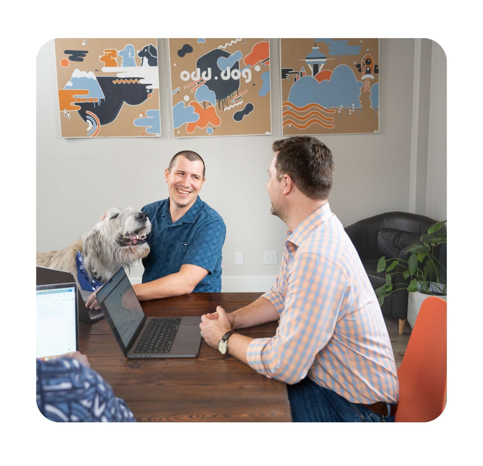 Two men — both with fair skin and brown hair — and an Irish Wolfhound smile at each other while seated at a table in front of an 