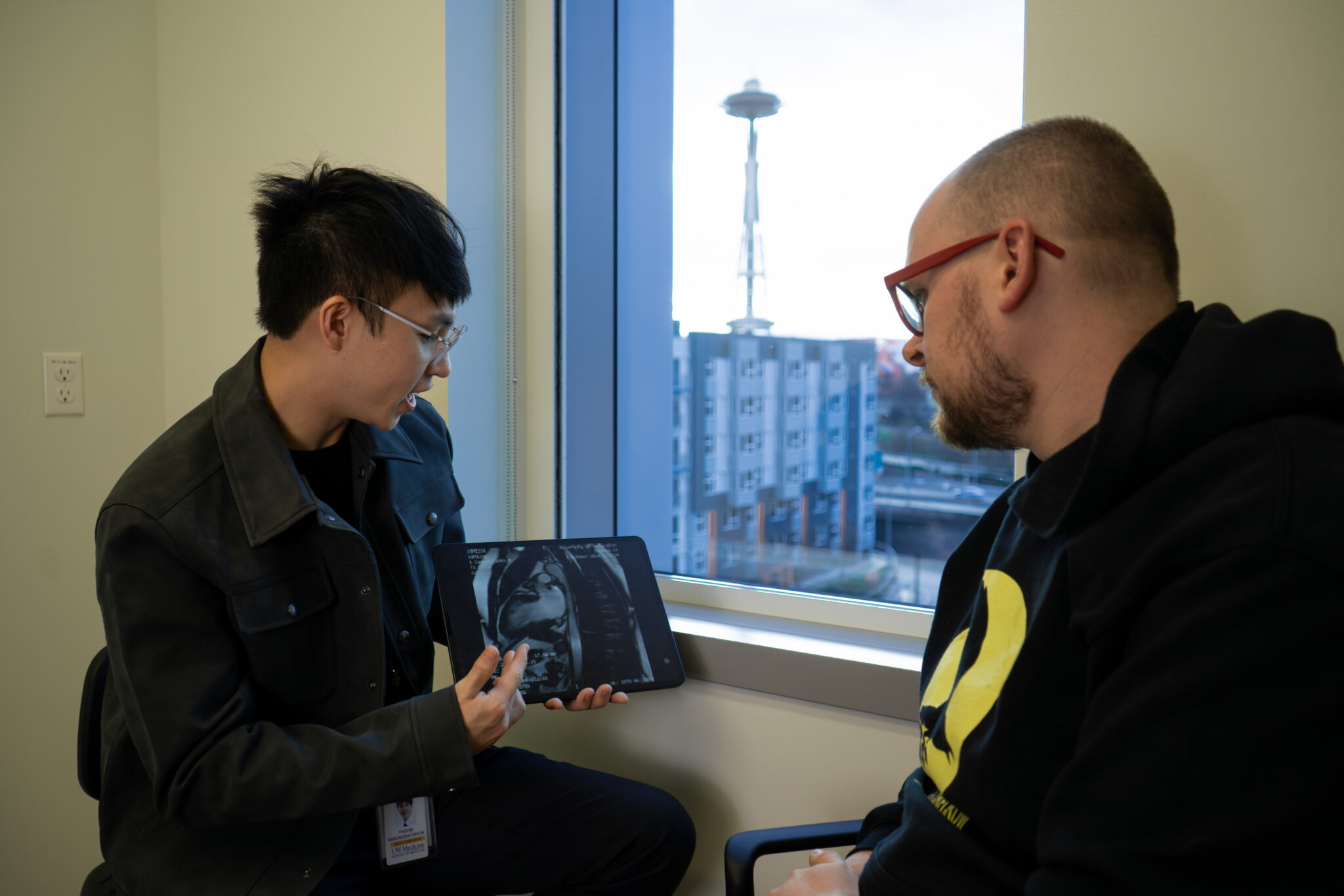 A UWMDI doctor shows a medical scan on a tablet to a patient sitting by a window, with the Seattle Space Needle visible outside.