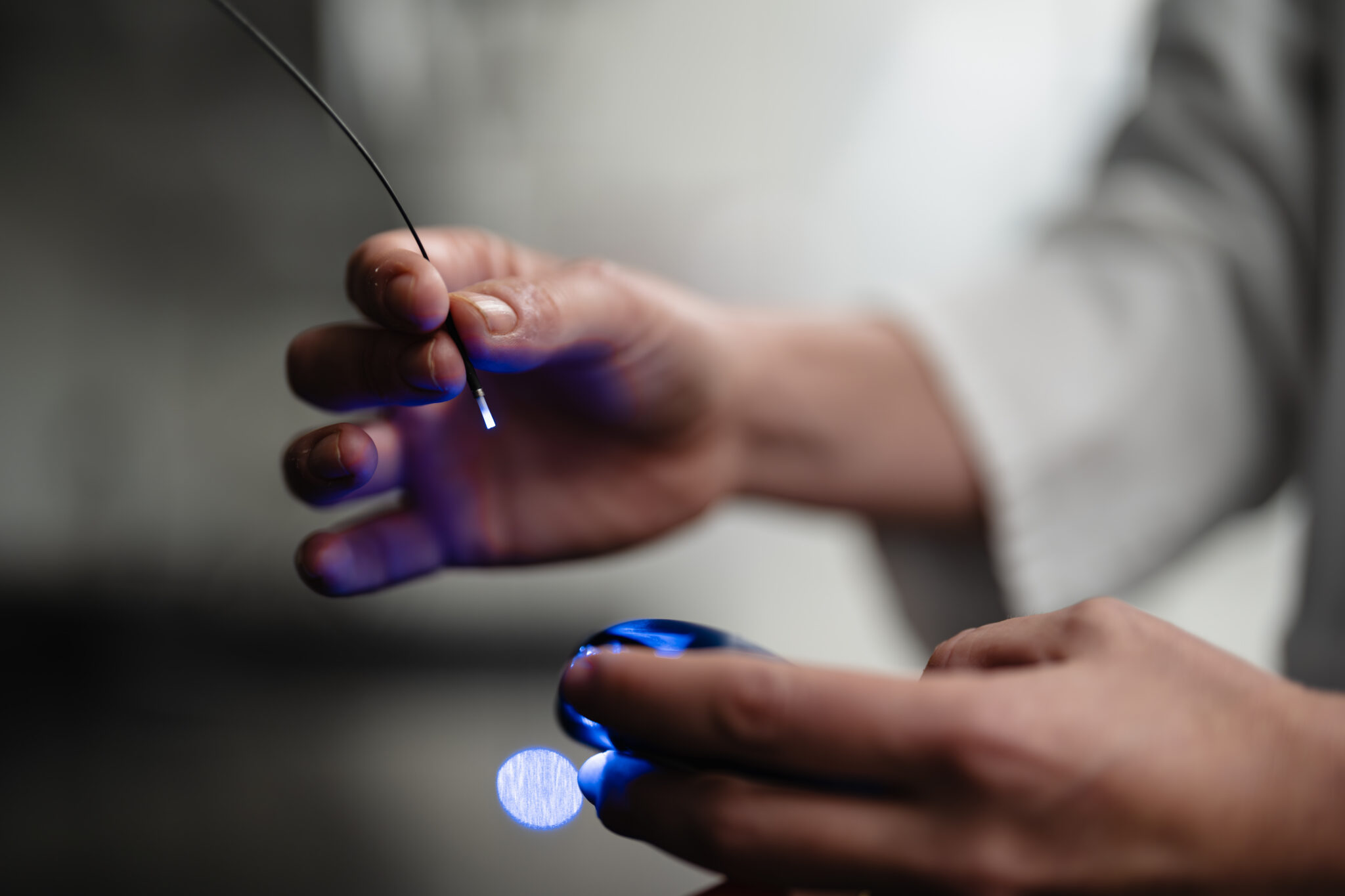 Close-up of a UWMDI researcher’s hands holding a thin fiber optic probe emitting blue light, examining it carefully during a lab experiment.
