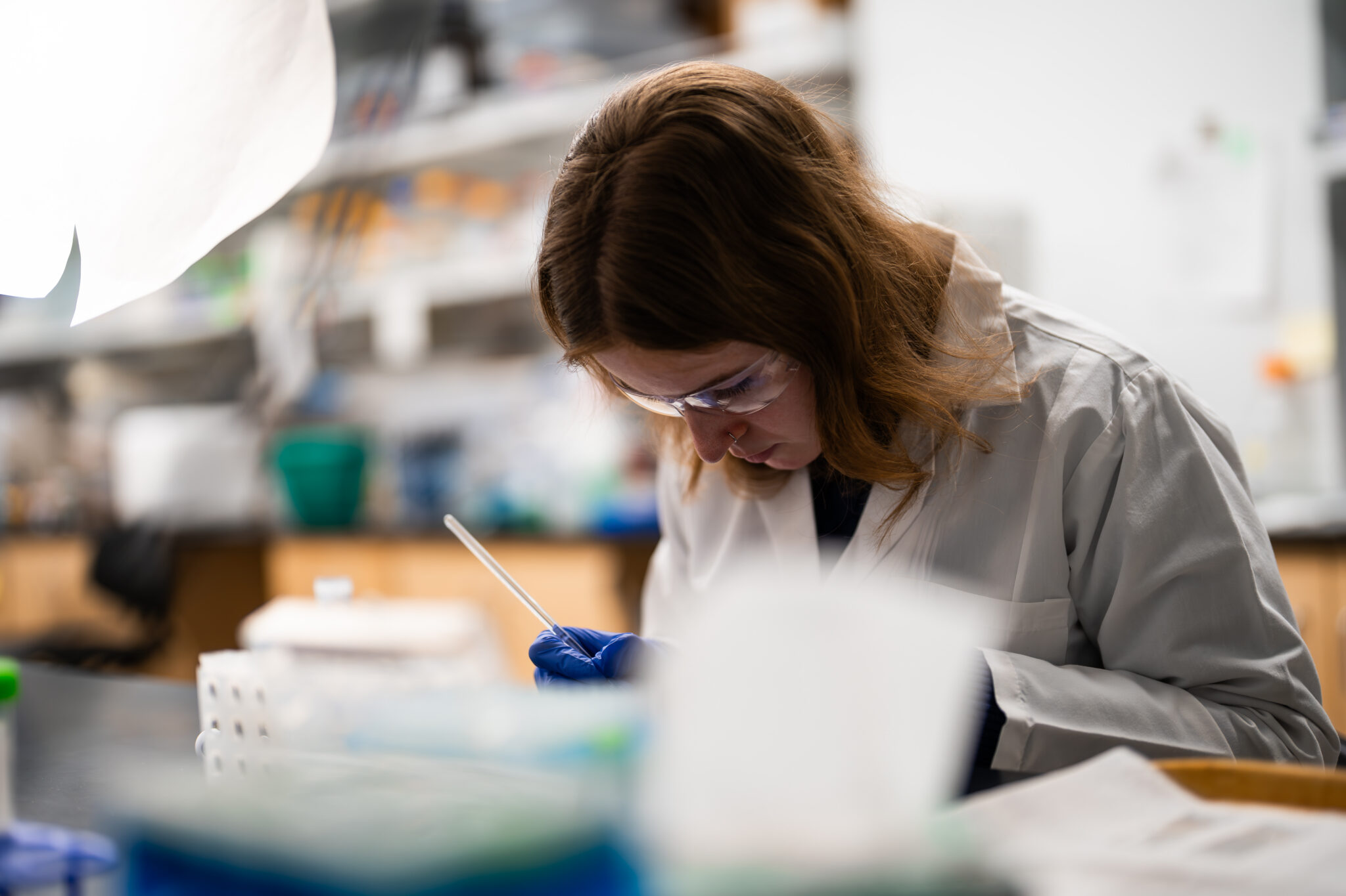 A person wearing a white lab coat, safety goggles, and gloves uses a pipette while working at a UWMDI laboratory bench, with blurred lab equipment and shelves in the background.