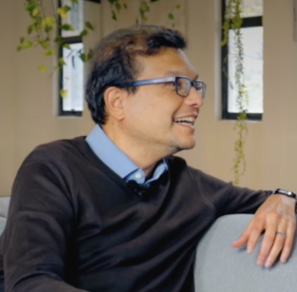 Man of Indian descent with glasses, collared shirt, and sweater smiles to his left