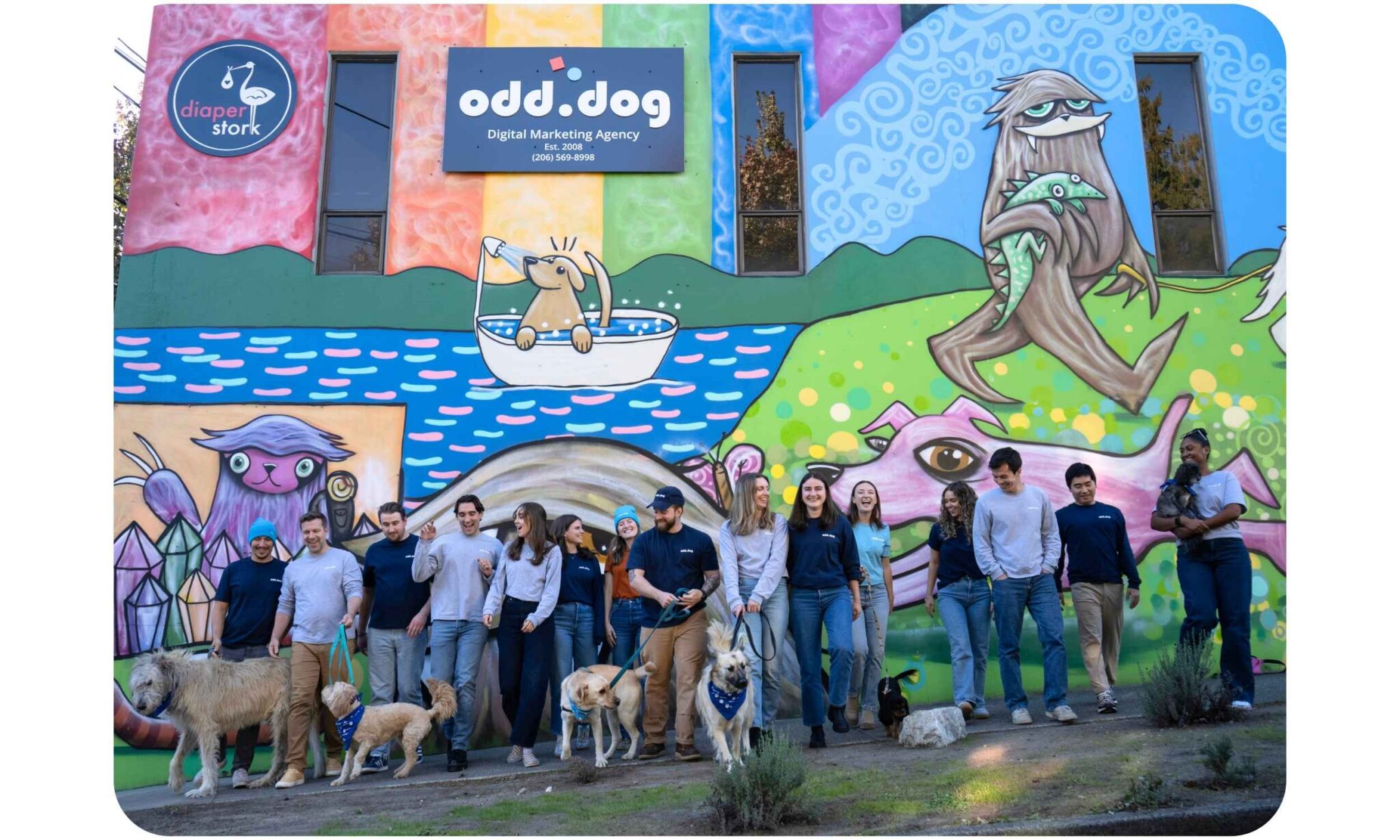 15 people and 5 dogs on leashes walk toward camera in front of colorful mural on building and Odd Dog sign