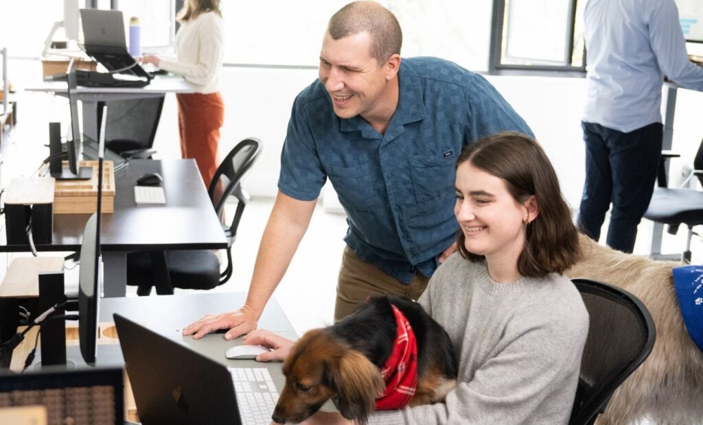 Two people smiling while looking at a laptop in an office. One person is seated with a dog wearing a red bandana, discussing how to make a website ADA compliant, while the other stands nearby. Coworkers work at standing desks in the background.