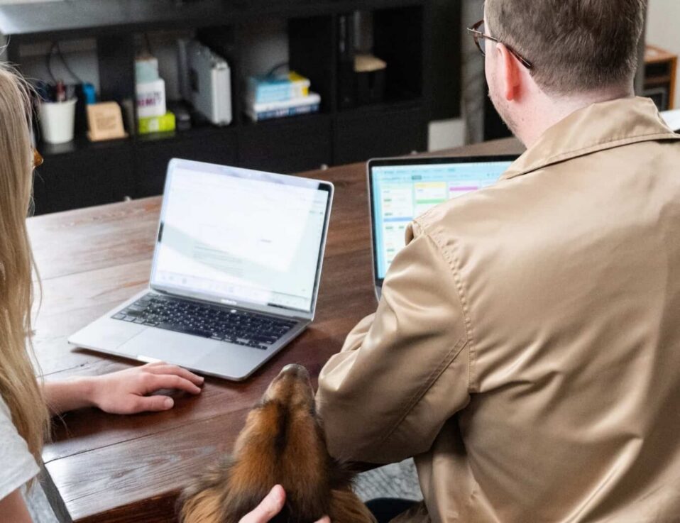 Two people sit at a wooden table working on laptops, with a brown dog resting its head on one person’s arm. In the background, a black TV stand holds books and small items—perhaps researching local services ads for their next project.