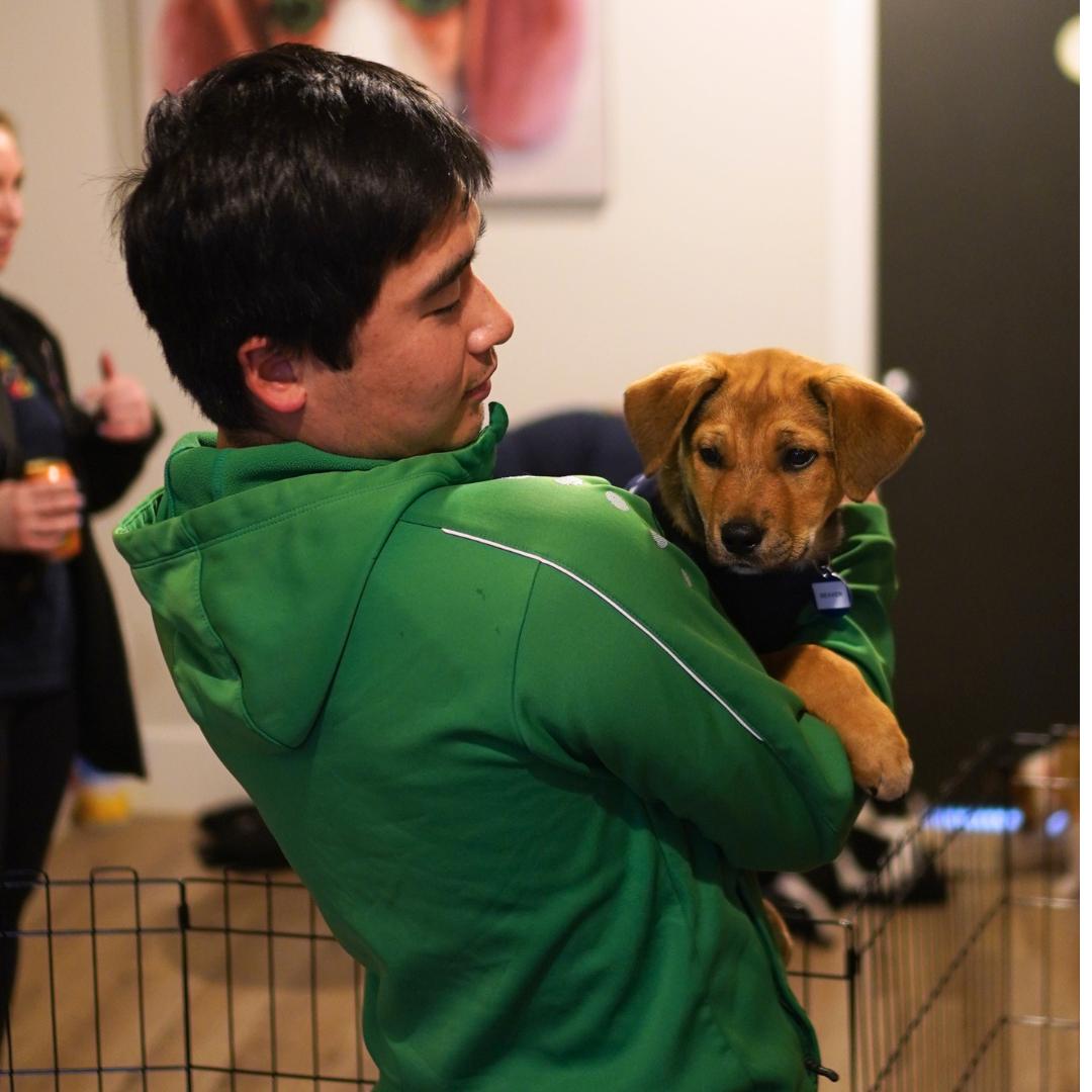 A man in a green jacket lovingly holds a brown puppy in his arms while standing indoors near a pet playpen. Another person is blurred in the background.