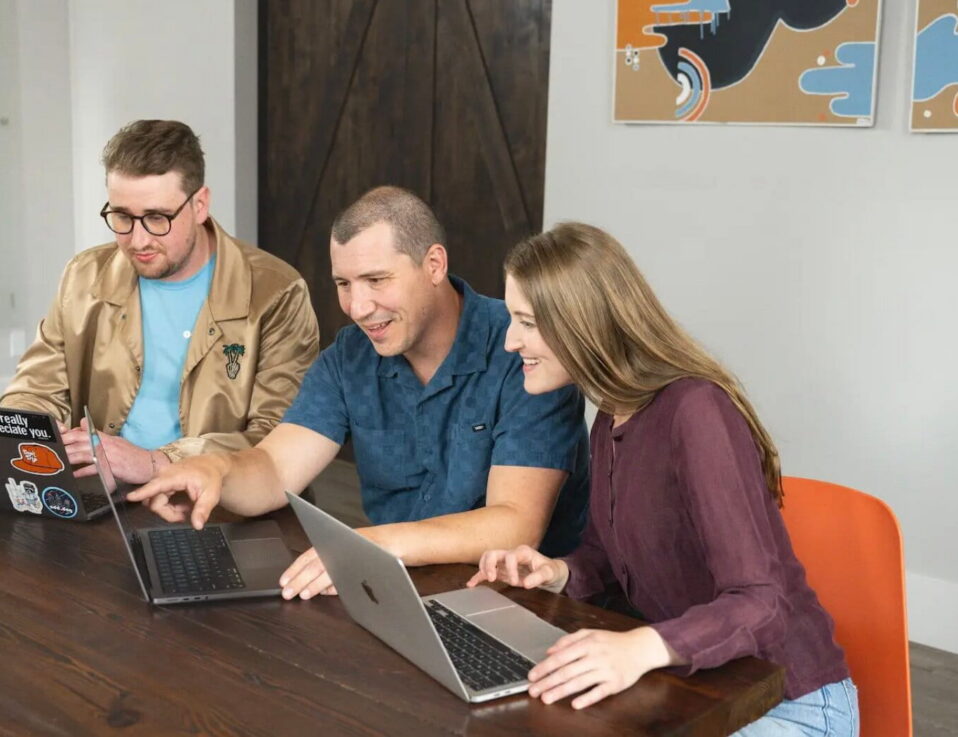 Three people sit at a wooden table, each using a laptop. They appear engaged and are smiling, discussing local services ads while one person points at a laptop screen. Two abstract art pieces hang on the wall behind them.