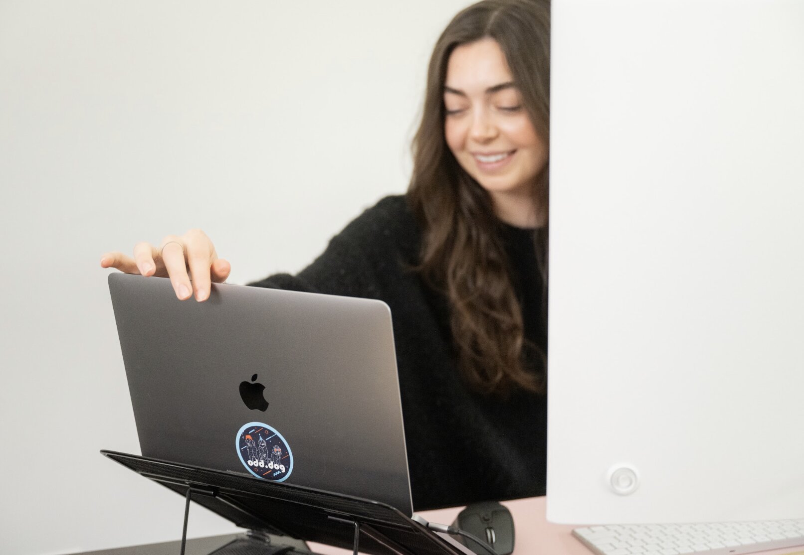 A woman with long brown hair smiles while sitting at a desk. She is closing a laptop with a sticker on it, possibly after researching how to make a website ADA compliant, next to a desktop monitor.