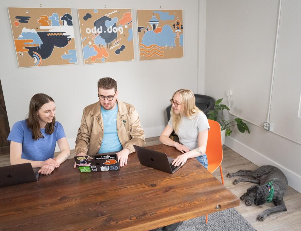 Three people sit at a wooden table with laptops, collaborating. Colorful artwork and the text ou.dog hang on the wall behind them. A black dog lies on the floor nearby. Sunlight comes in through a window.