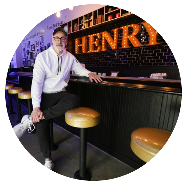 A person in glasses and a white hoodie leans on a bar counter with HENRY in large lit letters on the wall behind, surrounded by barstools in a diner-like setting.