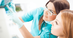 A dentist wearing blue gloves points to an x ray while explaining the results to a patient.