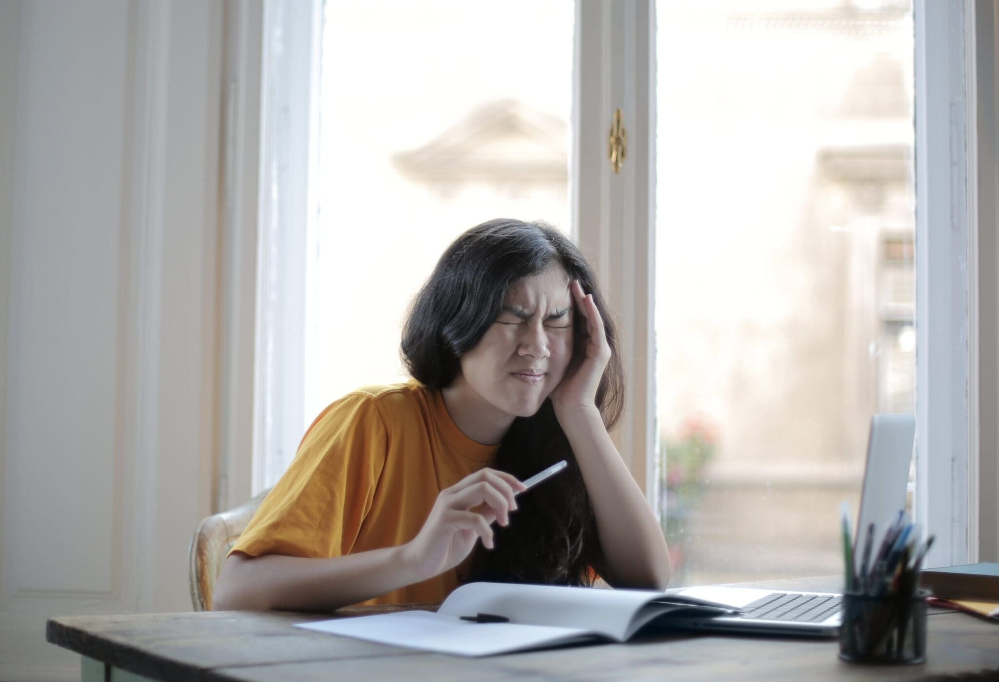A woman having a migraine or headache, sitting at a desk, working on a laptop, and holding a pen.