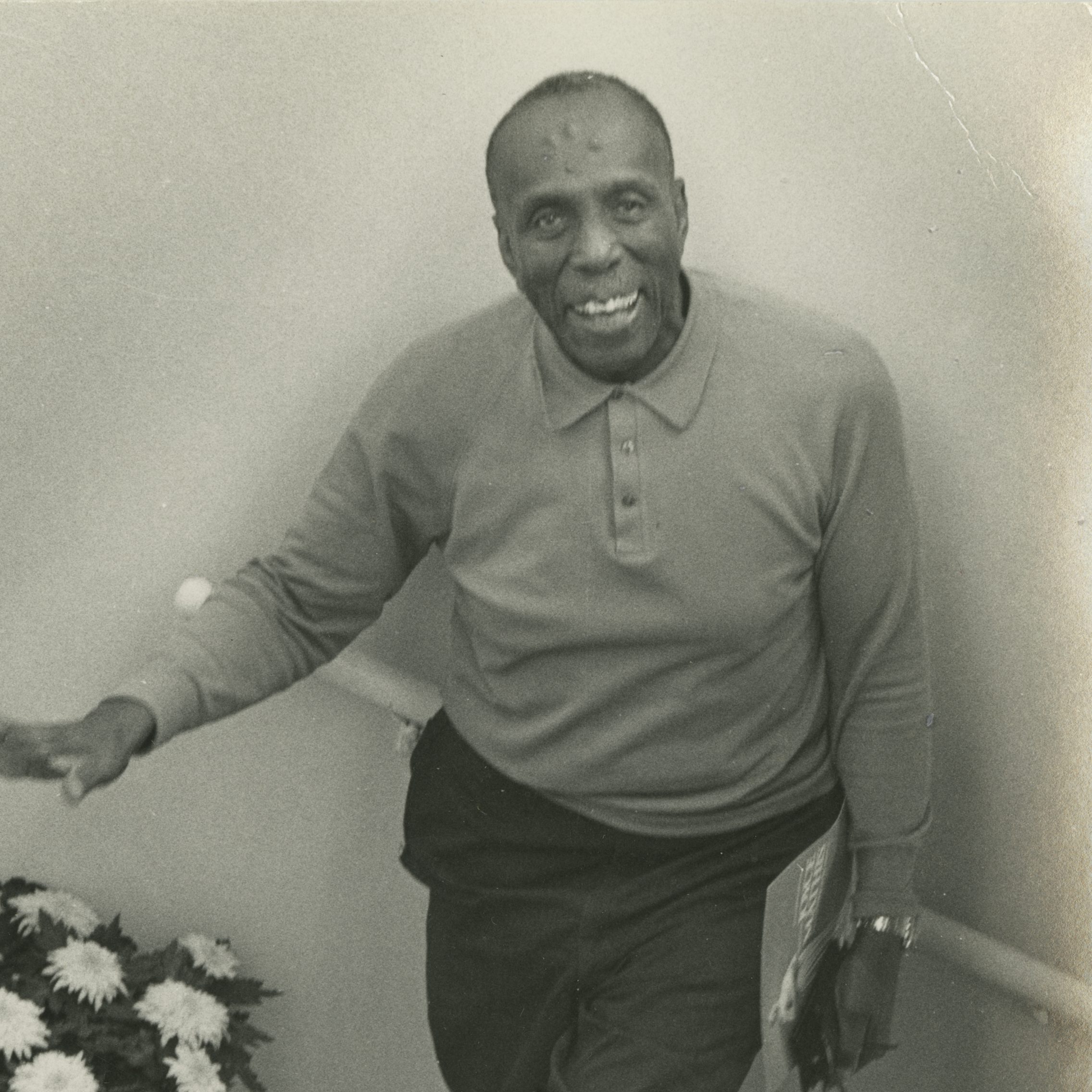 A black and white photograph of Howard Thurman smiling and standing in a stairwell. He holds his glasses and a sheet of paper in his left hand and gestures with his right hand. There is a pot of flowers in the stairwell, also.