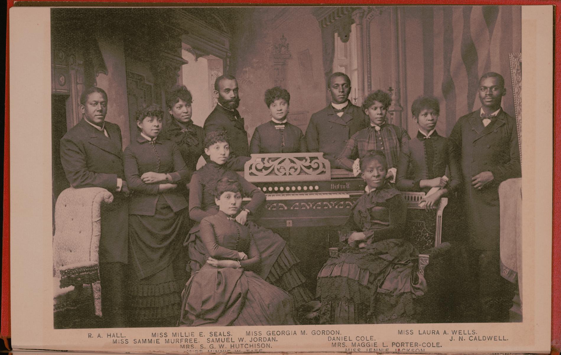 Sepia photograph depicting 12 of the Jubilee Singers dressed in clothing of the period. Three of them are seated in front of a piano, and the rest stand behind or beside it. The names listed at the bottom of the image are the following: R. A. Hall, Miss Millie E. Seals, Miss Georgia M. Gordon, Miss Laura A. Wells, Miss Sammie Murfee, Samuel W. Jordan, Daniel Cole, J.N. Caldwell, Mrs. S. G. W. Hutchinson, Mrs. Maggie L. Porter-Cole, Miss Minnie W. Tate, and Miss Jennie Jackson.