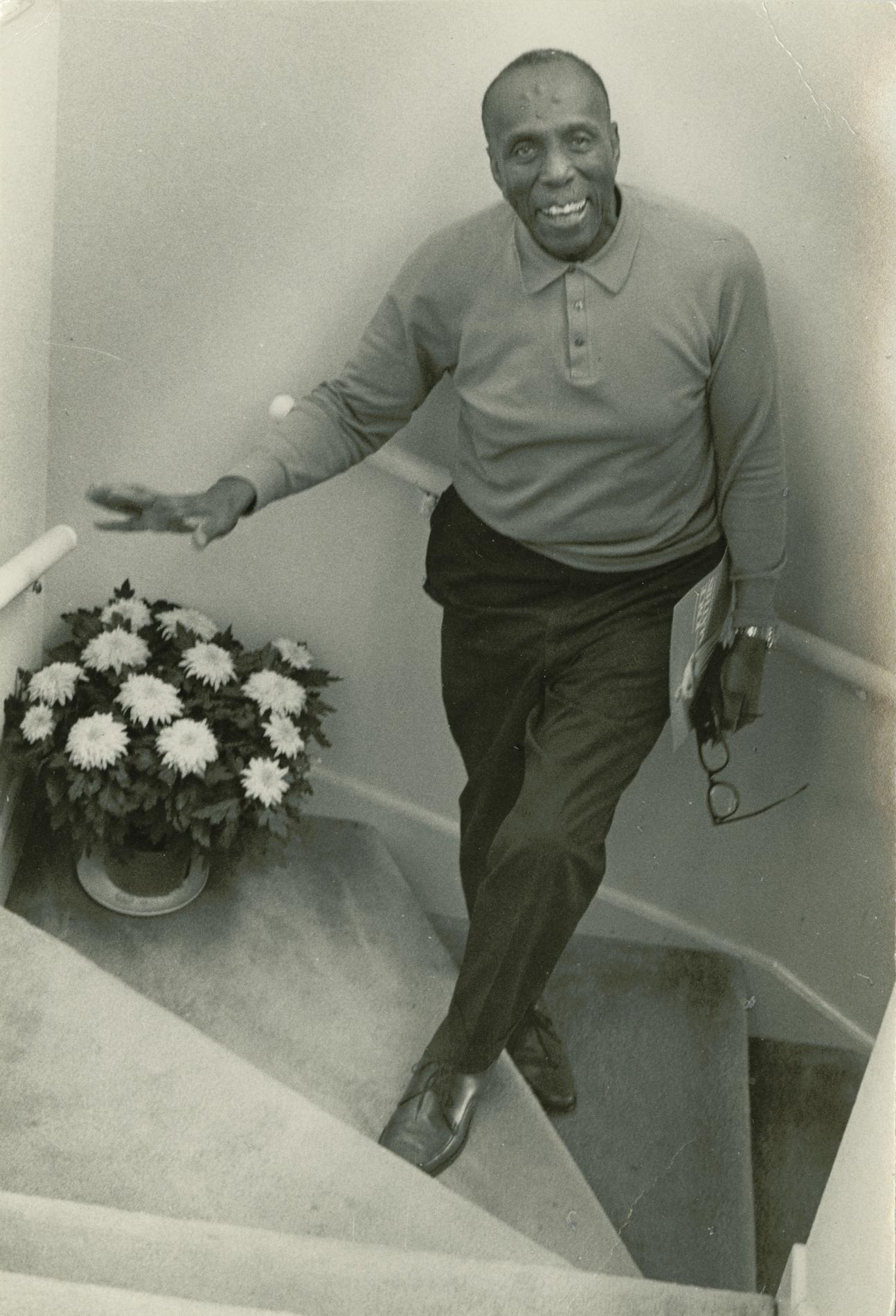 A black and white photograph of Howard Thurman smiling and standing in a stairwell. He holds his glasses and a sheet of paper in his left hand and gestures with his right hand. There is a pot of flowers in the stairwell, also.