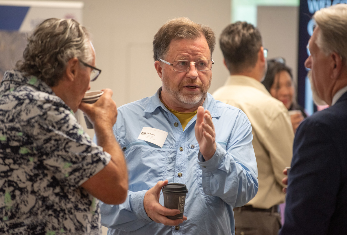 A man in a blue shirt talks to two other men drinking coffee.