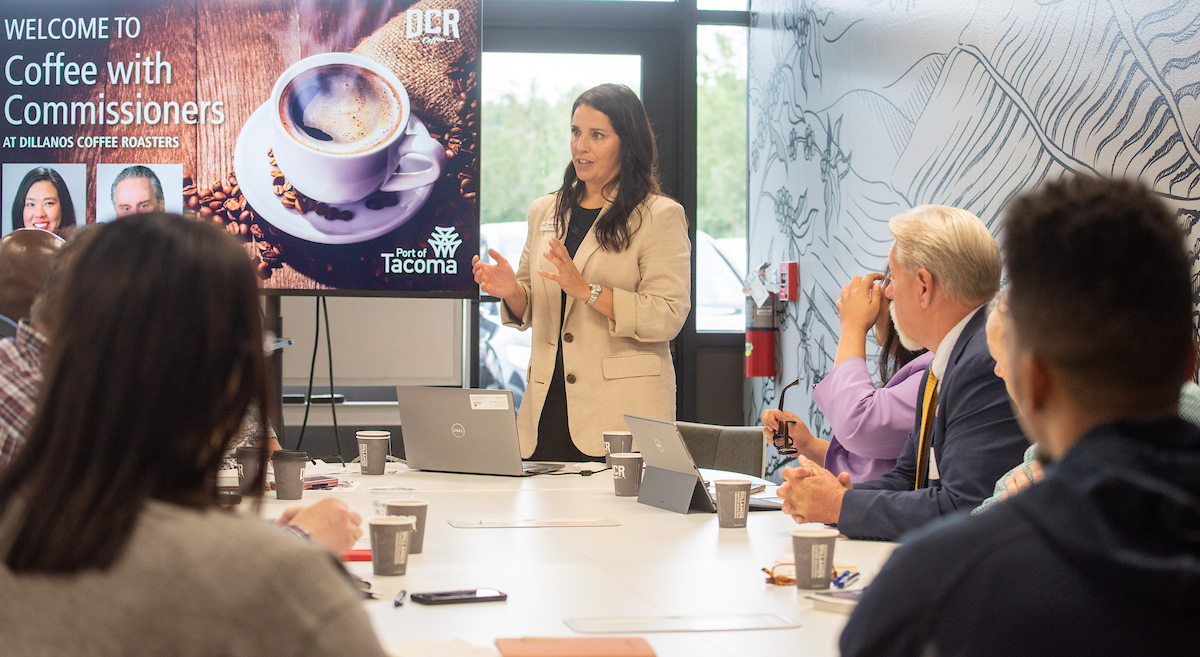A woman in a beige jacket talks to a group of people sitting down at a table with coffee.