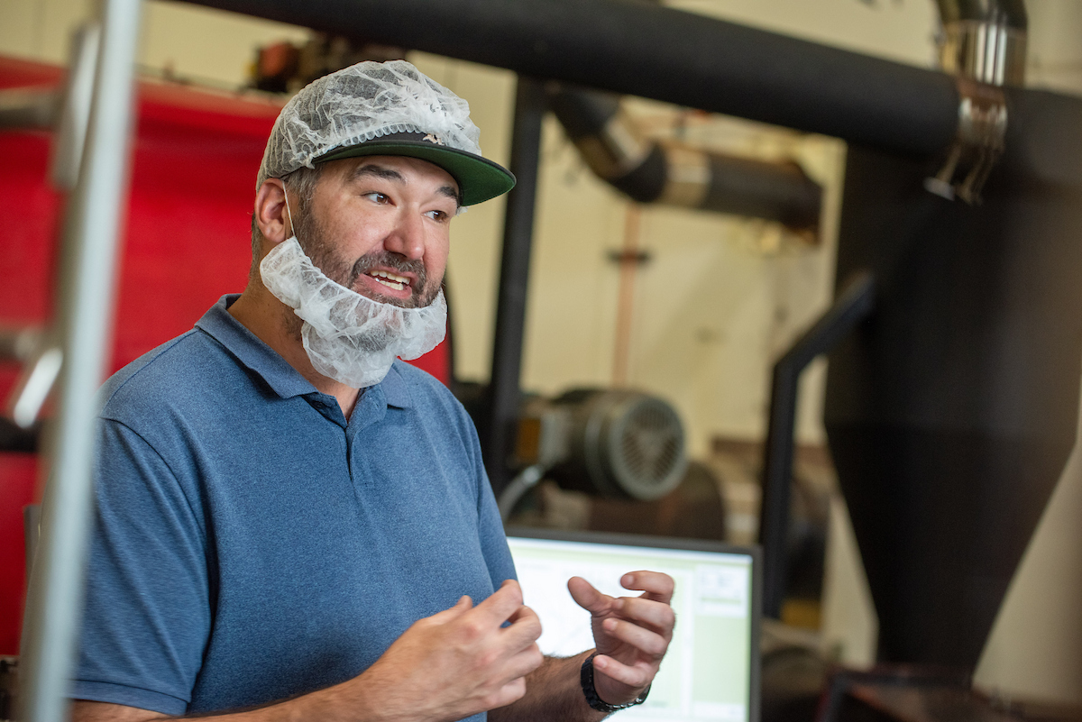 A man with a hair and beard net in a blue shirt explains something to someone off camera.