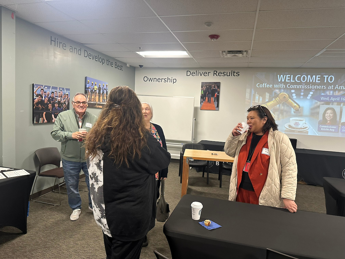 A woman in a white sweater drinks coffee while talking to a woman near a black table.