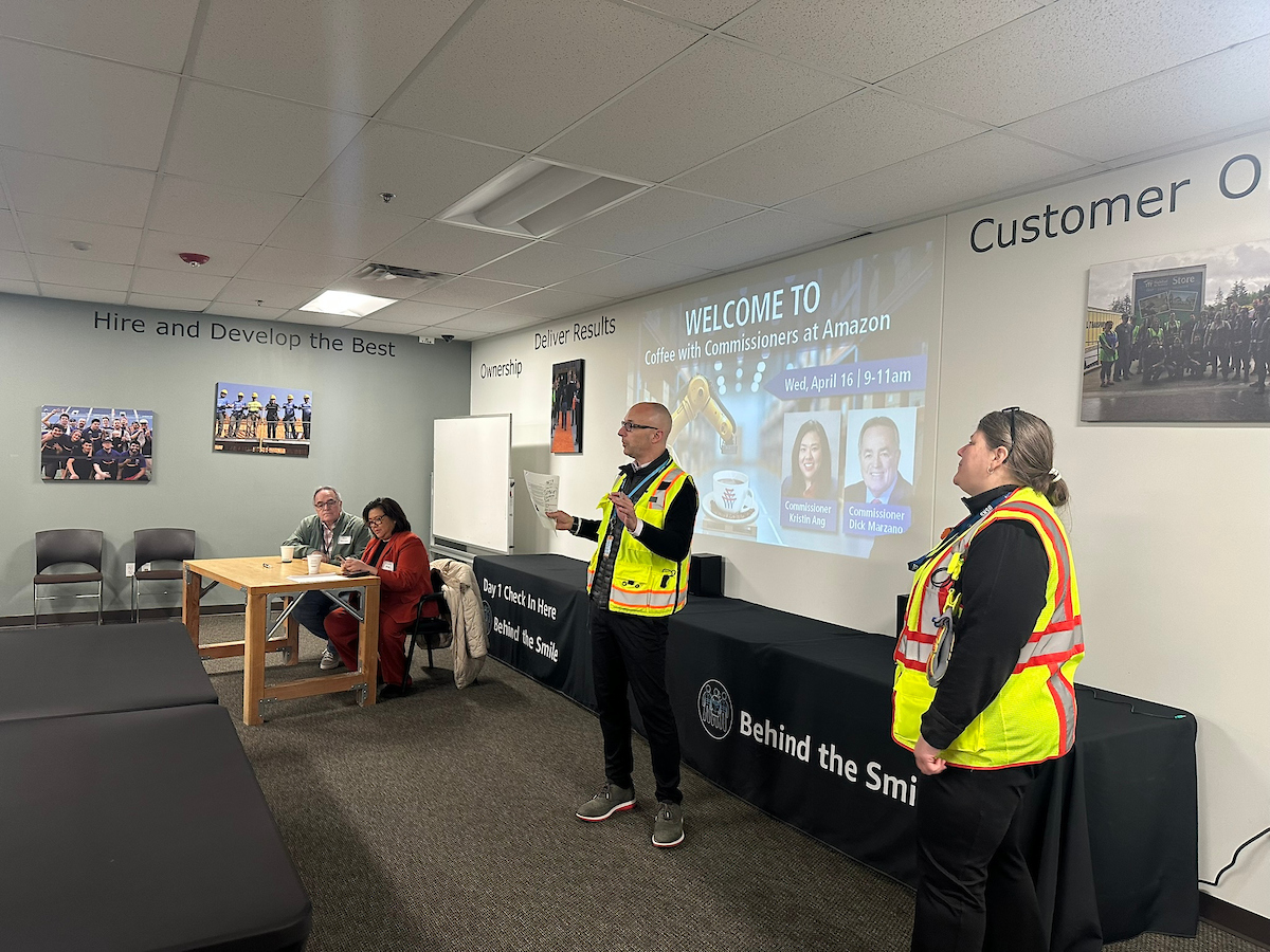 A man and woman in reflective vests stand in the front of a room giving a talk in front of a projector.