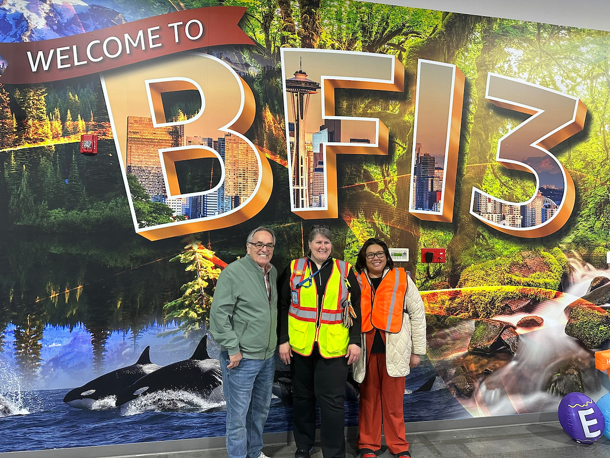 Two women and a man stand in front of a "Welcome to BF13" sign.