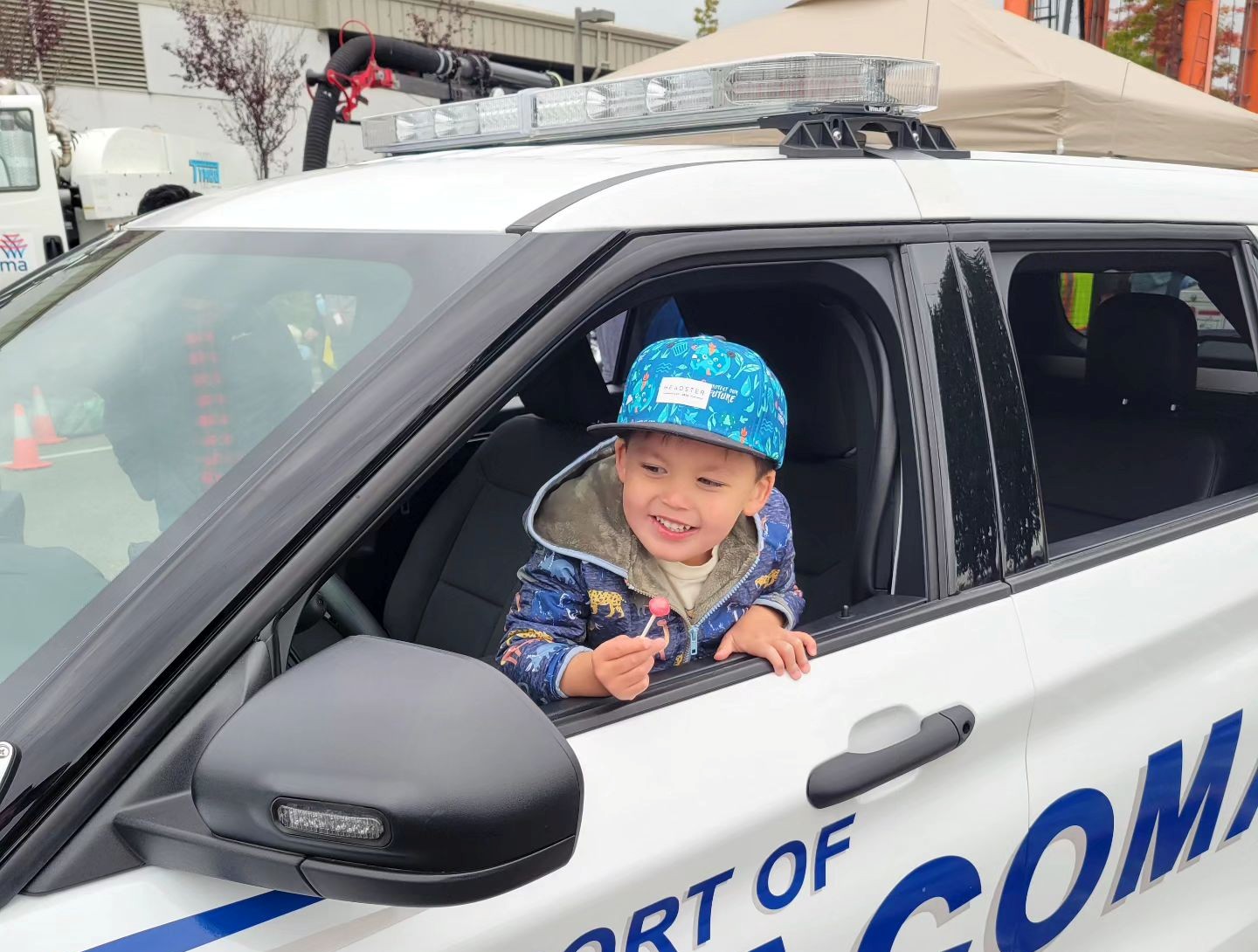A young boy in a blue cap smiles in the drivers seat of a patrol car.