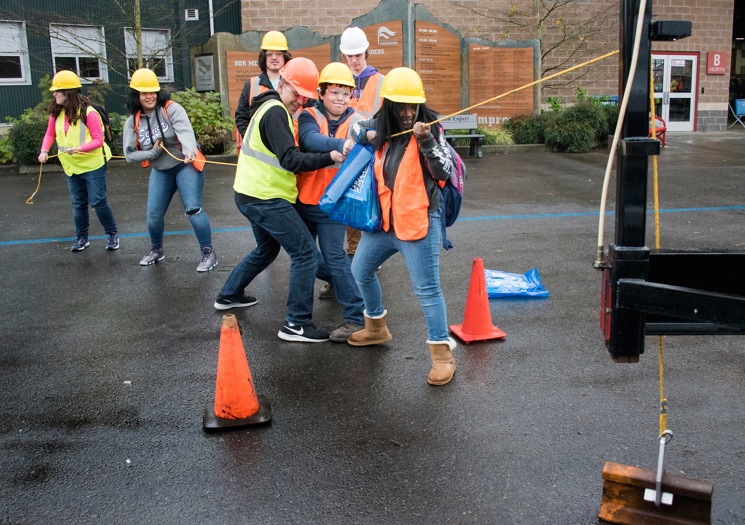 High schoolers in safety vests and hard hats work together to pull a rope on pavement.