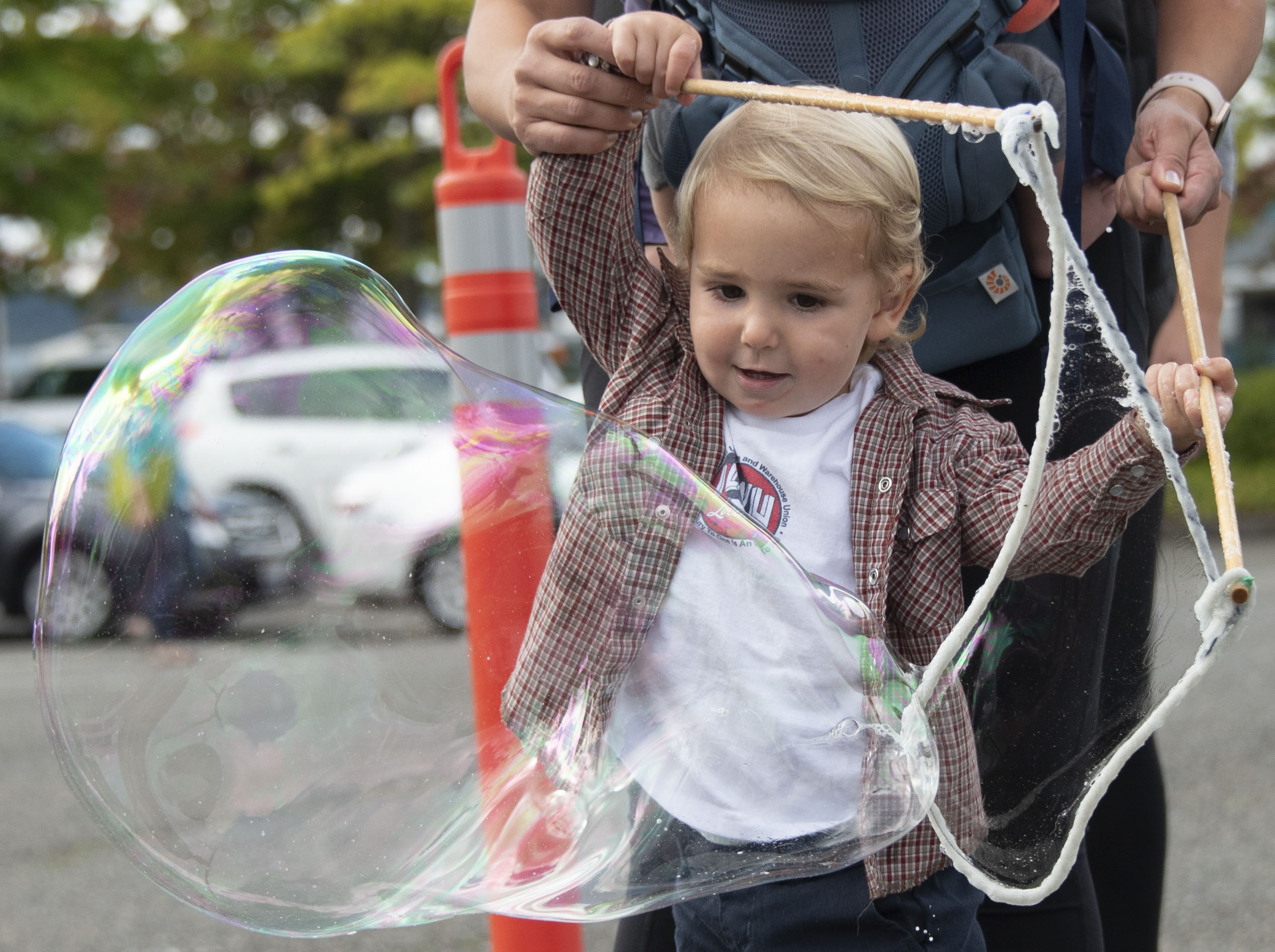 A toddler with blonde hair is assisted by an adult in making a huge bubble.