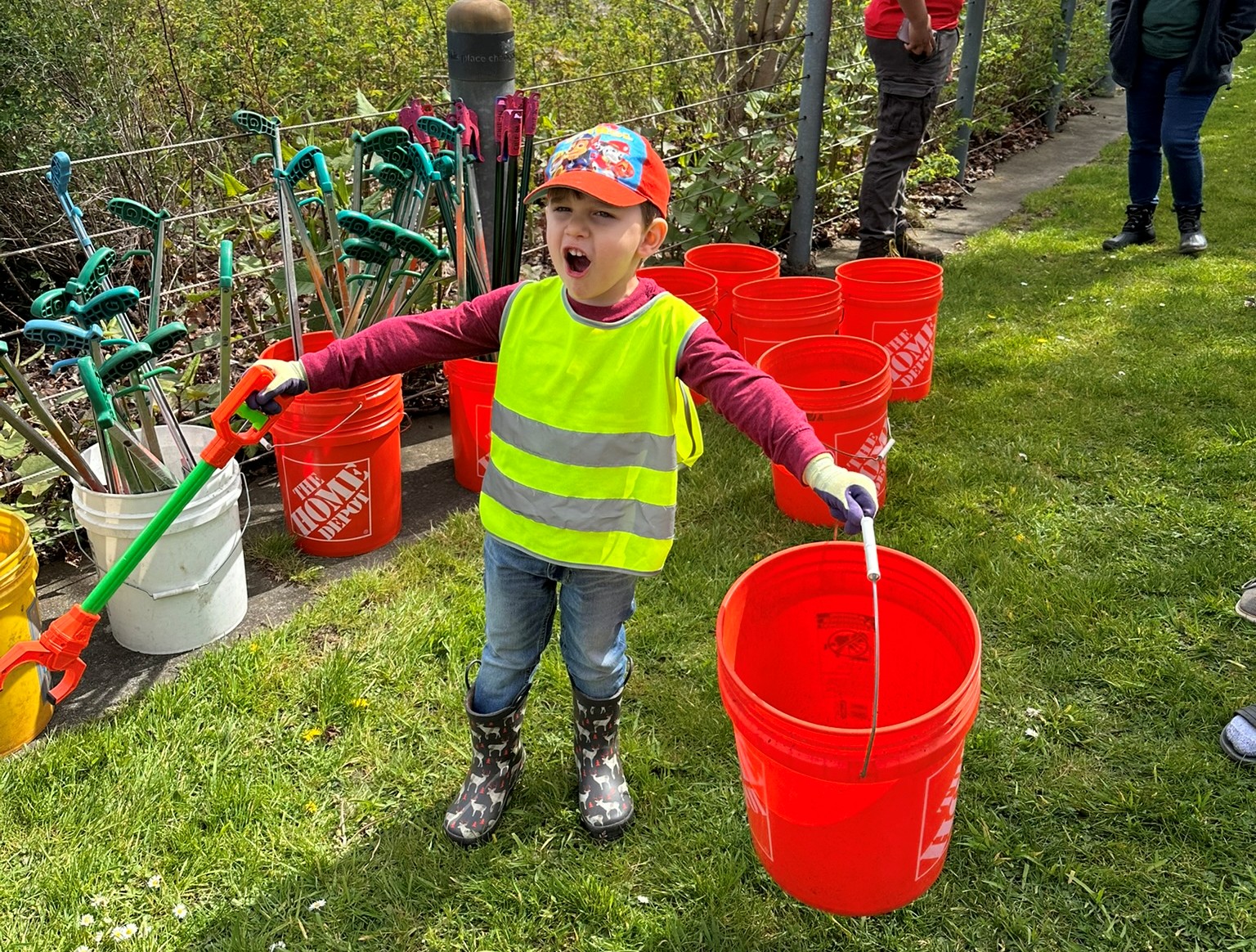 A young boy in safety geat holds an orange bucket with his hands outstretched 