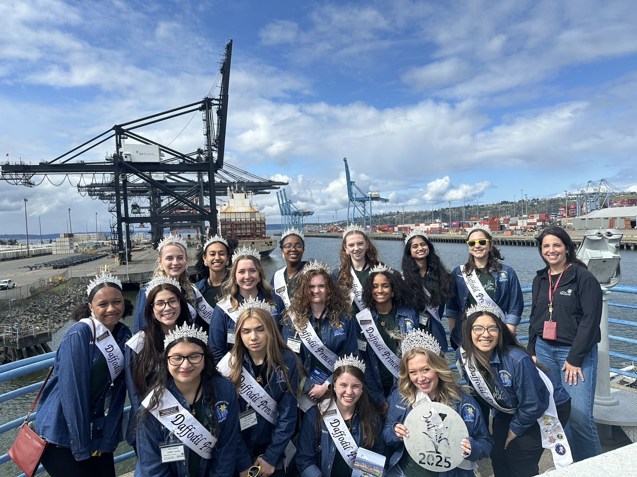 A group of young ladies in blue shirts and sashes pose in front of a cargo terminal.