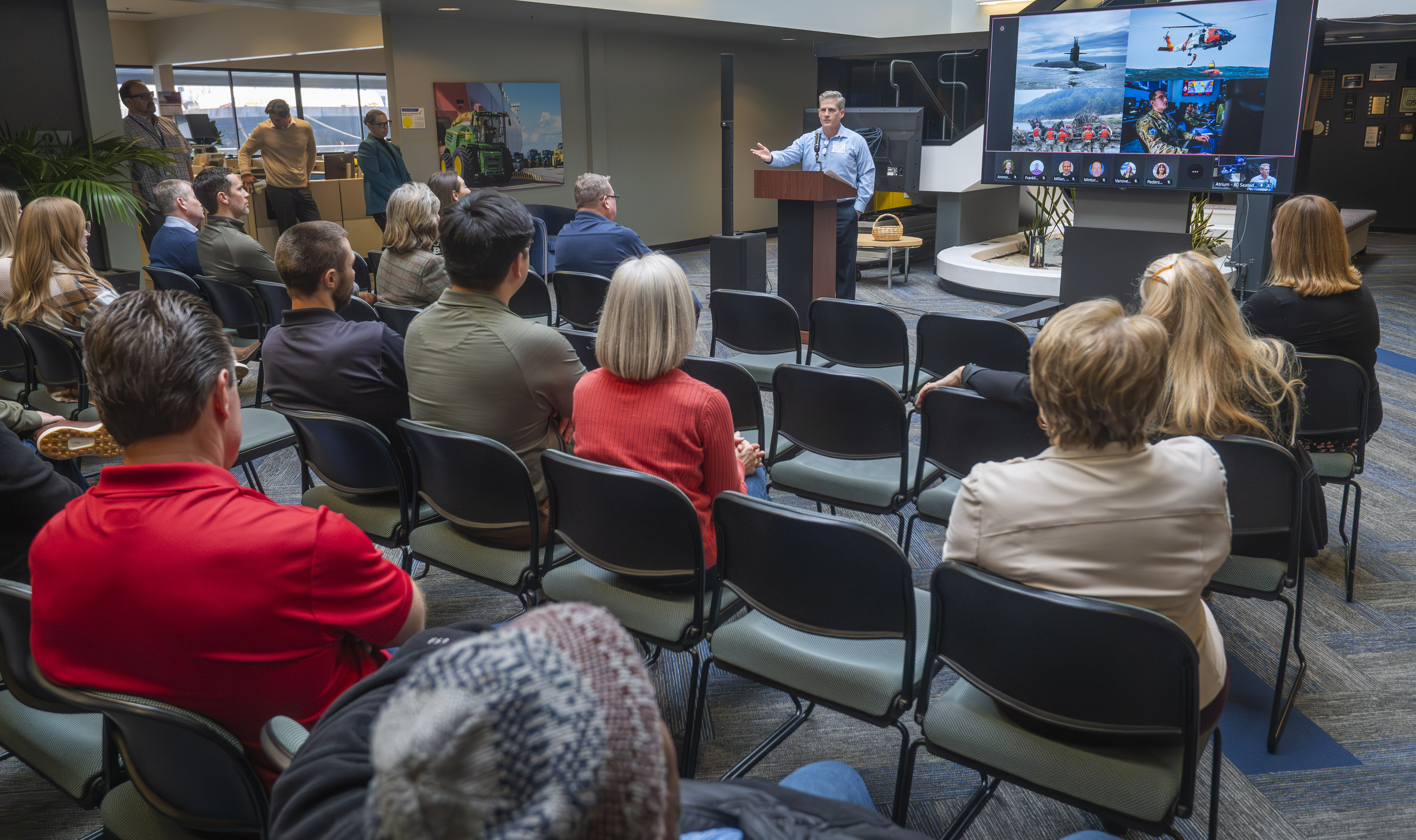 A speaker gestures to an audience in front of a screen