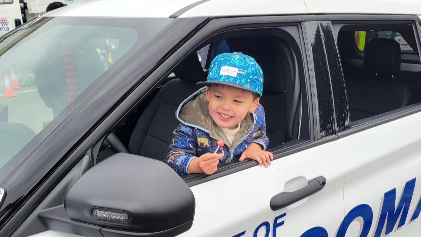 A young boy in a blue cap smiles while leaning out the window of a security vehicle. 
