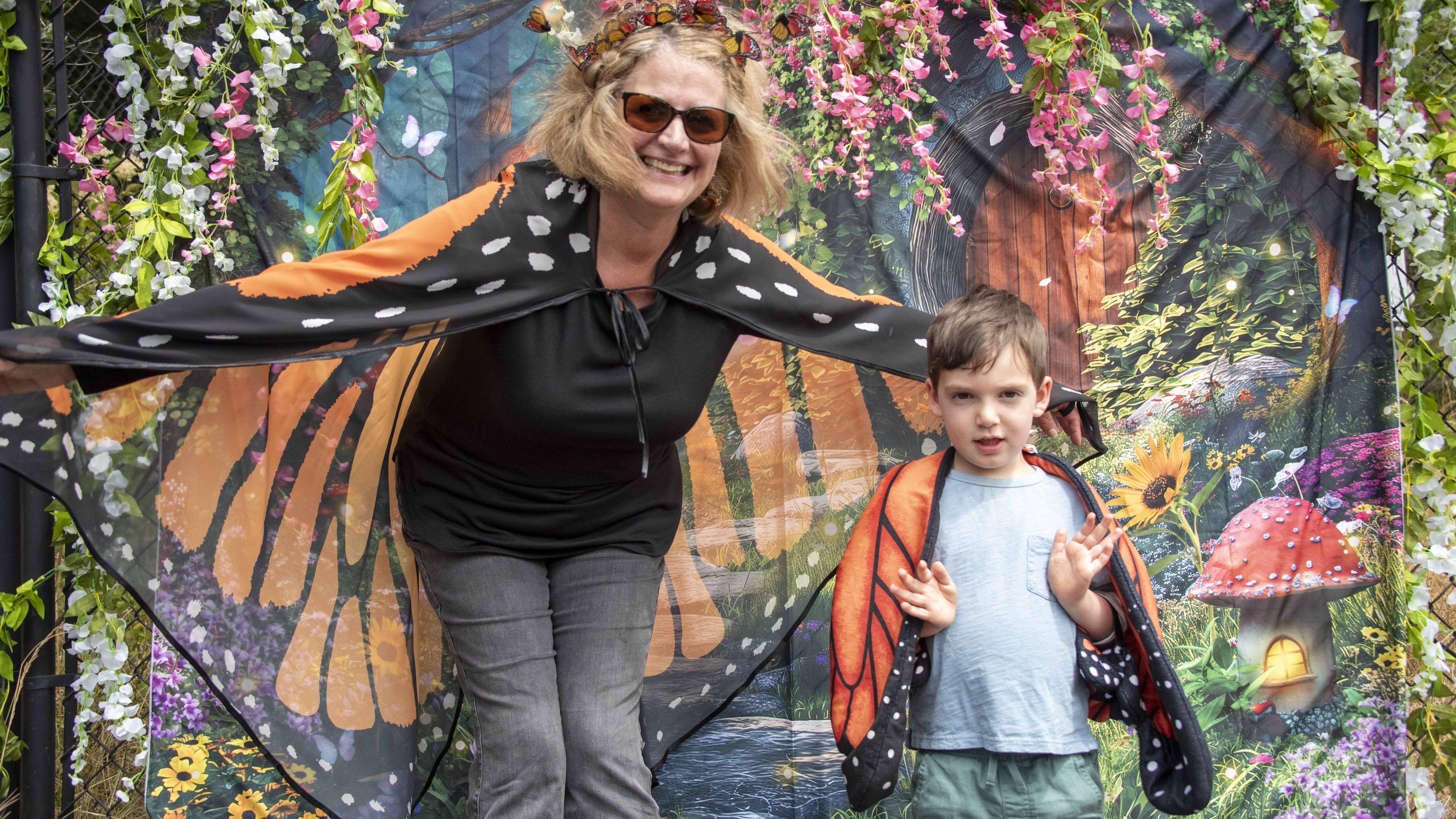A woman and young boy wearing butterfly wings in front of a fairy photo backdrop.