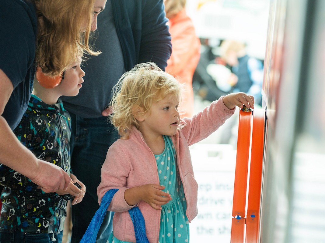 A small girl in a pink coat moves a toy car on an orange track. 