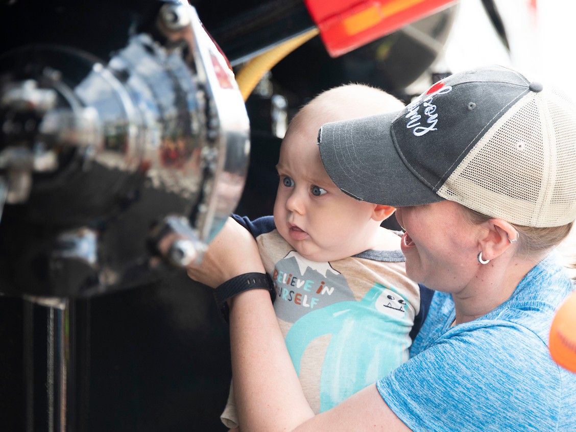 A caregiver smiles while holding their baby up to a big truck. 