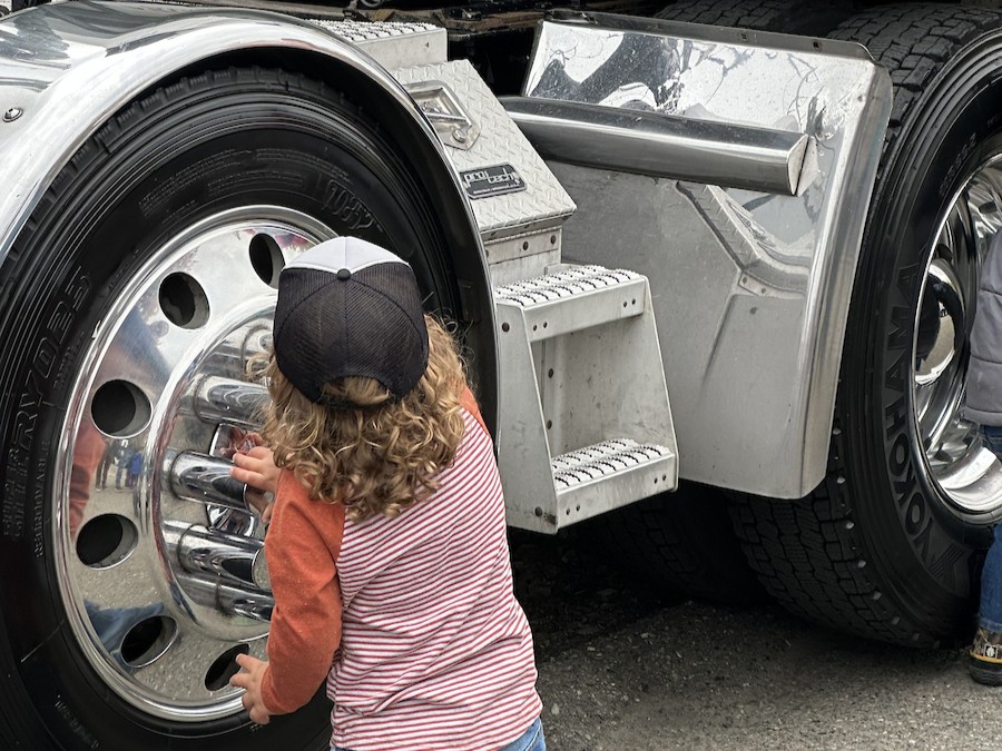 A young child in a baseball cap touches the rims of a large truck.