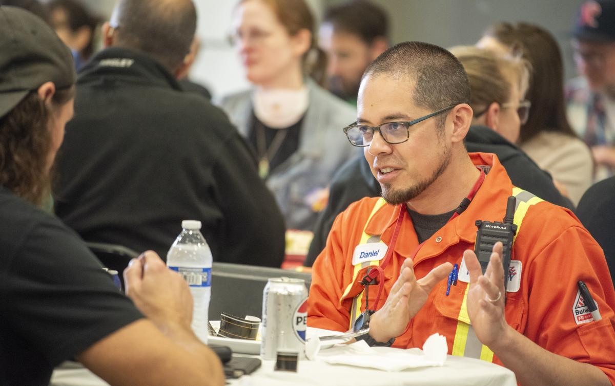 A man wearing glasses and high-visibility coveralls speaks to another man at a lunch table.