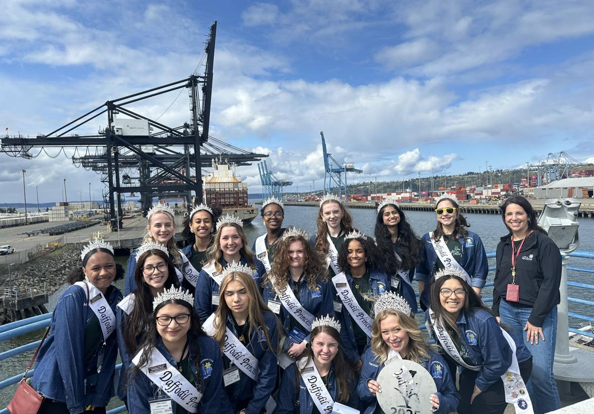 A group of young ladies in blue shirts and sashes pose in front of a cargo terminal.