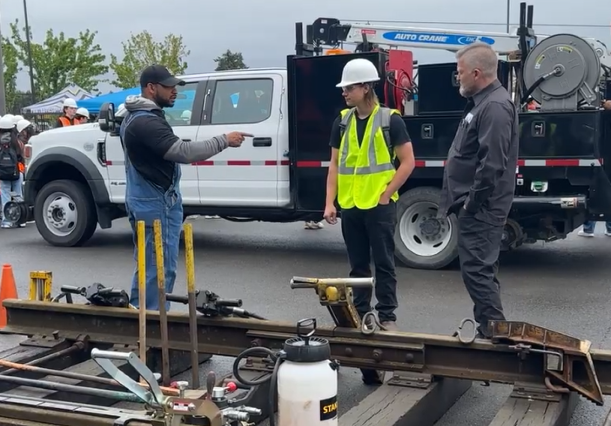 Two men at a rail track demonstration speak to a student in safety gear.