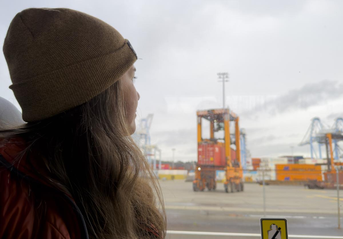 A woman with long hair wearing a beanie looks out a bus window at a port terminal
