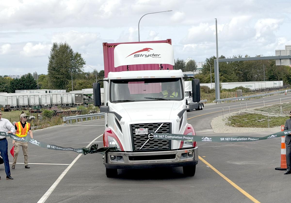 A semi truck drives through a ribbon to celebrate the opening of SR167 expressway as community partners hold the ribbon.