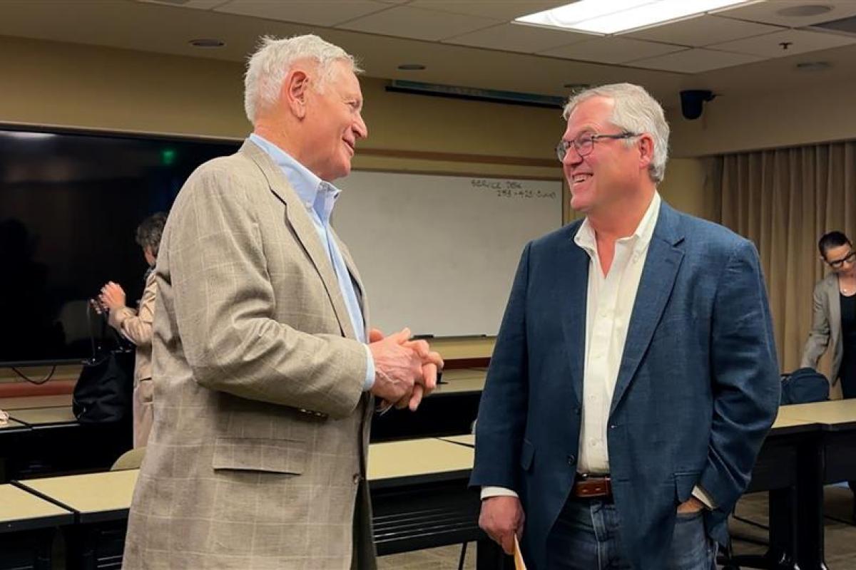 Two older men smile at each other at a public meeting.