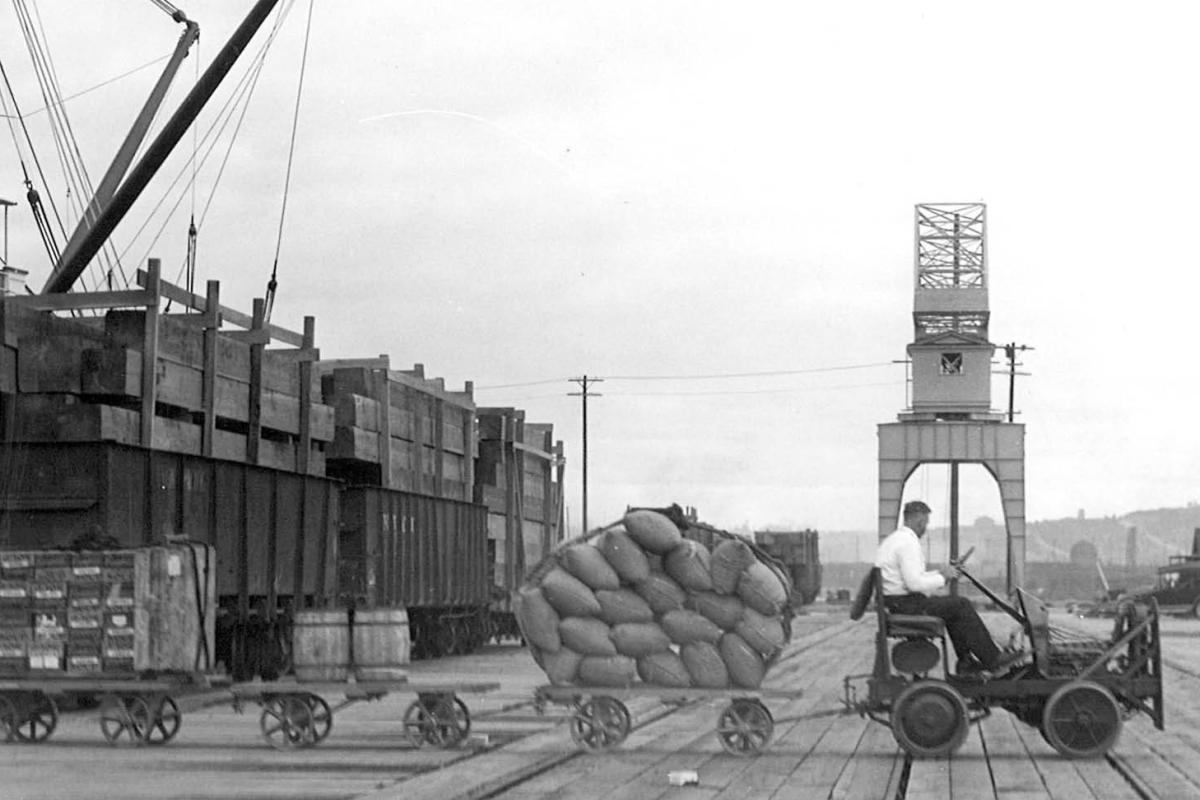 Black and white photo of a man pulling a line of carts on a port terminal.