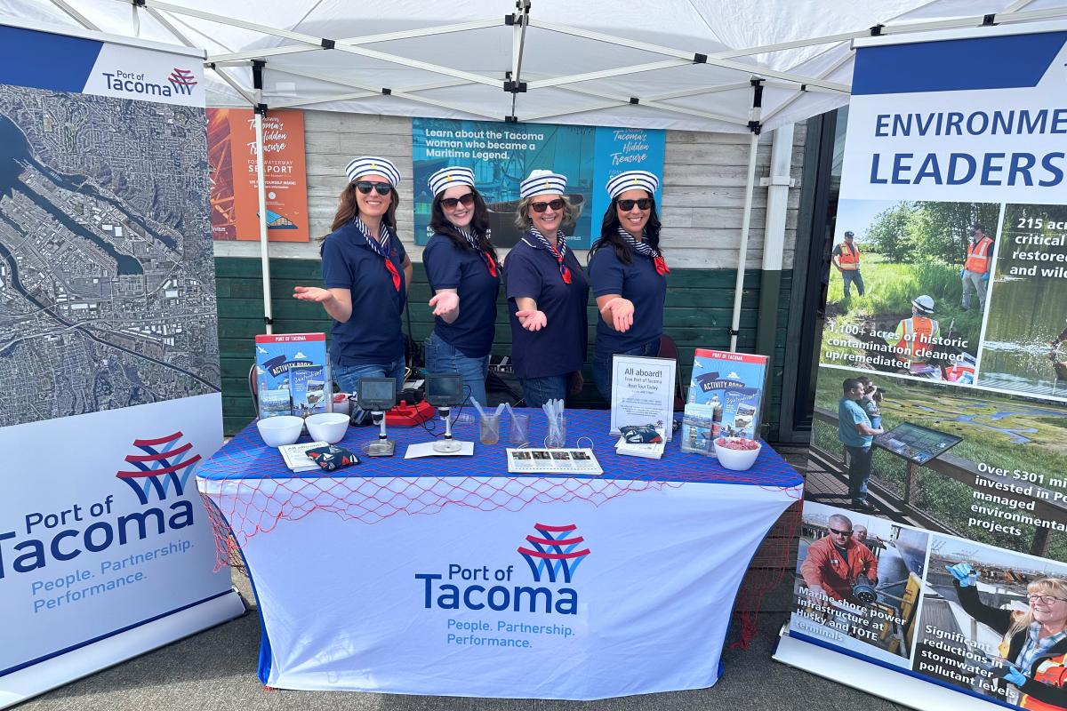 Four women in sailor hats hold their hands out while standing behind a Port booth at Oceanfest 2024.
