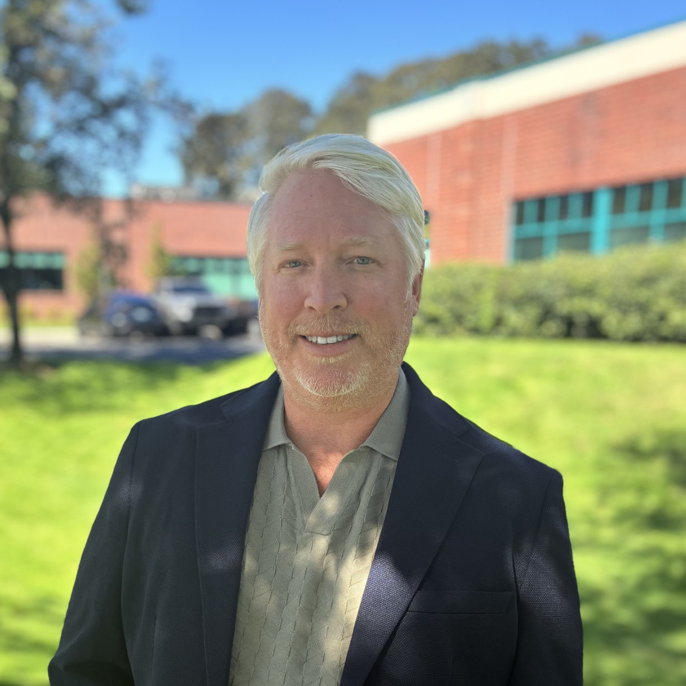 Headshot of an employee named Steve with a blue sky, green grass, trees and brick building in the background